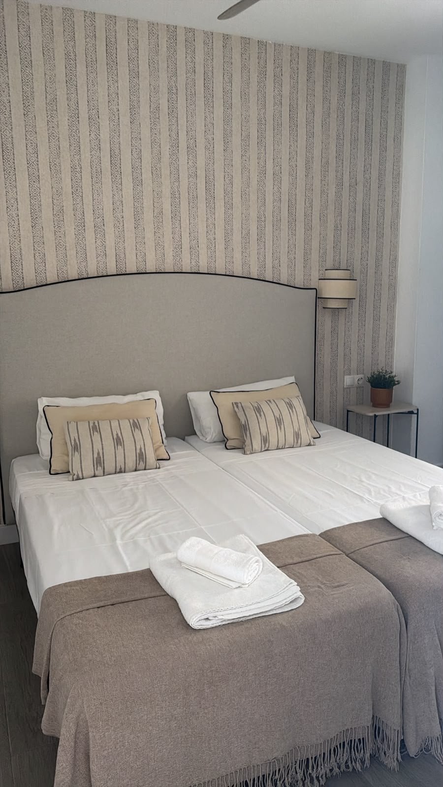 Modern bedroom with striped wallpaper, gray upholstered headboard, white bedding, decorative pillows, and folded towels on the foot of the bed
