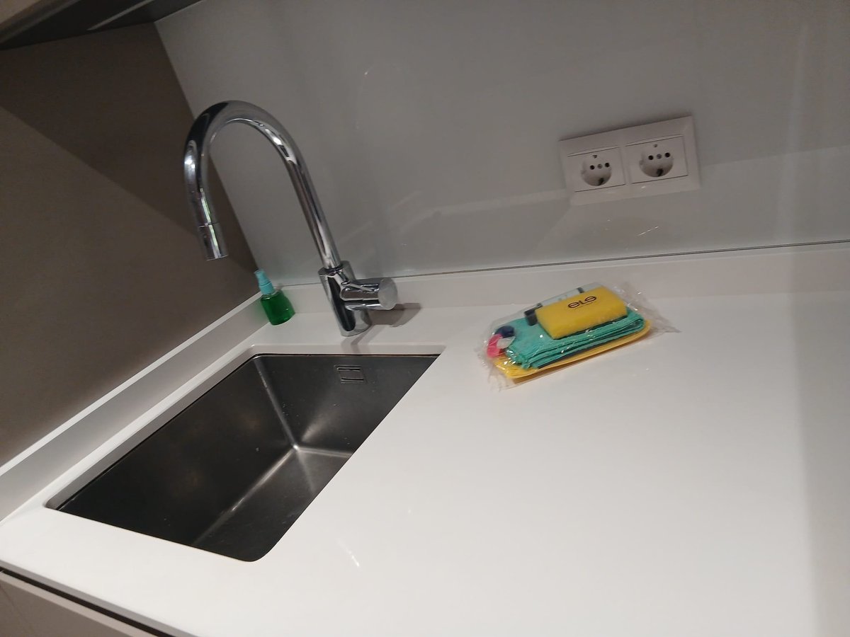 Modern kitchen sink with chrome faucet and green aerator, stainless steel basin, white countertop with sponges, electrical outlets visible on backsplash