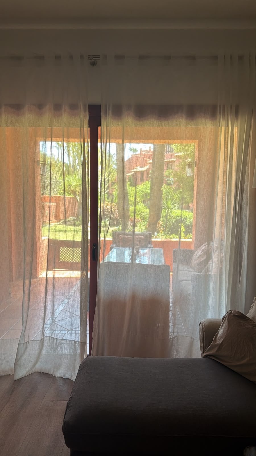 Bright sunlit room with sheer curtains, glass doors opening to a brick patio and garden with green plants