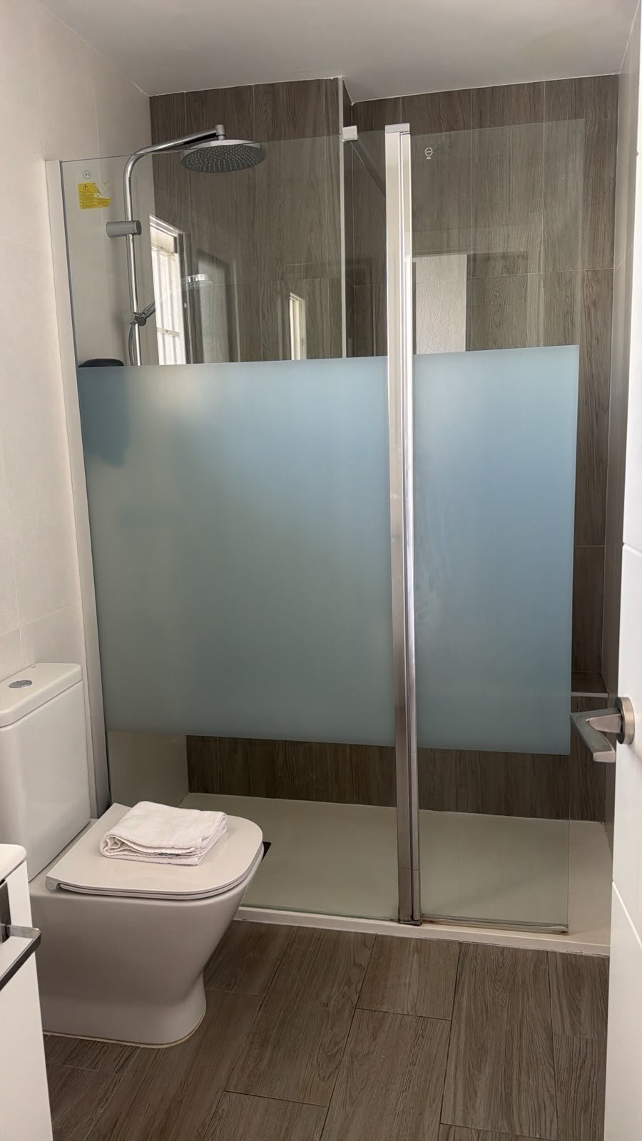 Modern bathroom with frosted glass shower enclosure, rain showerhead, dark wood tile walls, white toilet, and light wood-look flooring