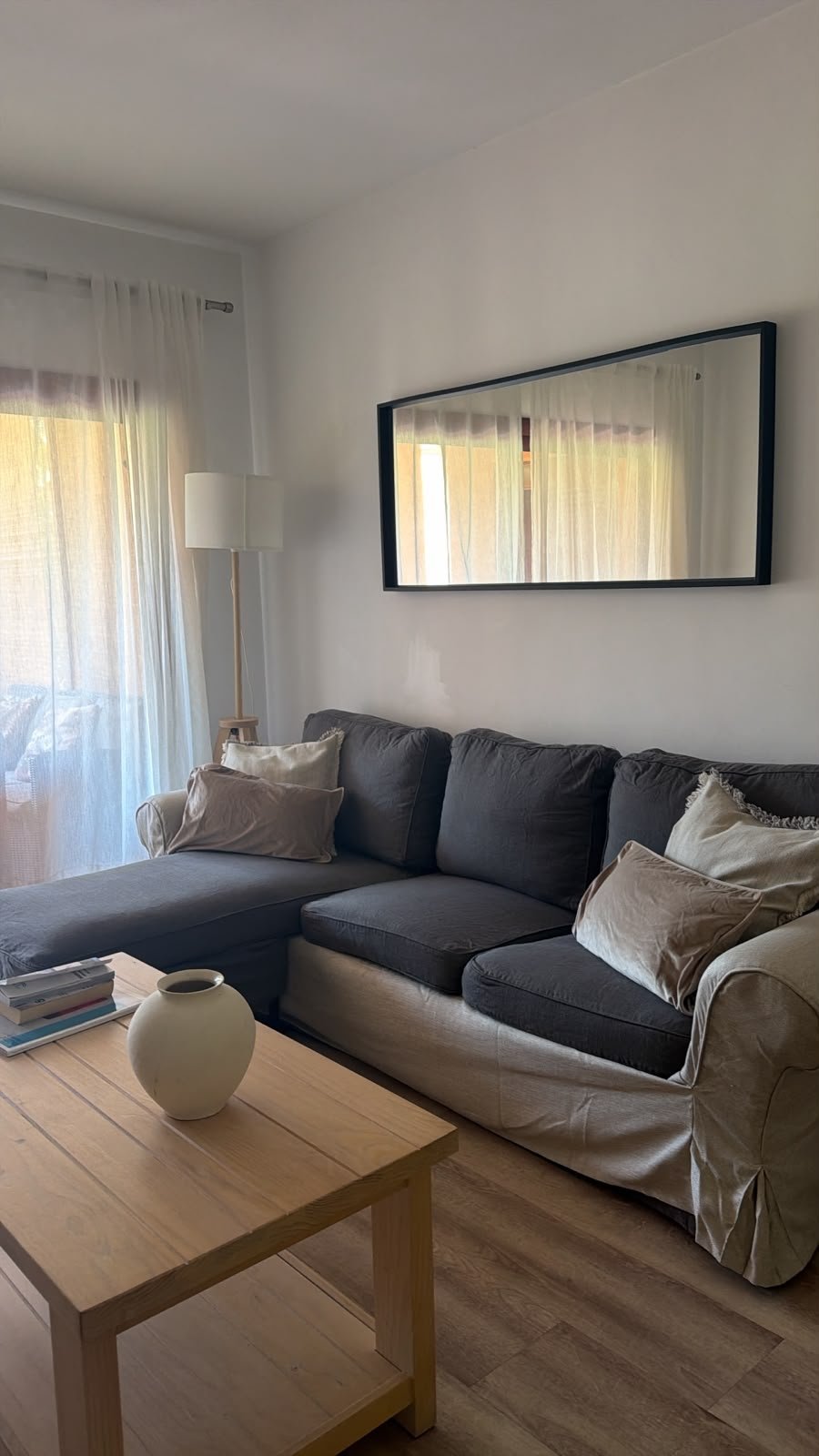 Modern living room with dark and light two-tone sectional sofa, wooden coffee table, mirror on white wall, and sheer curtains by window
